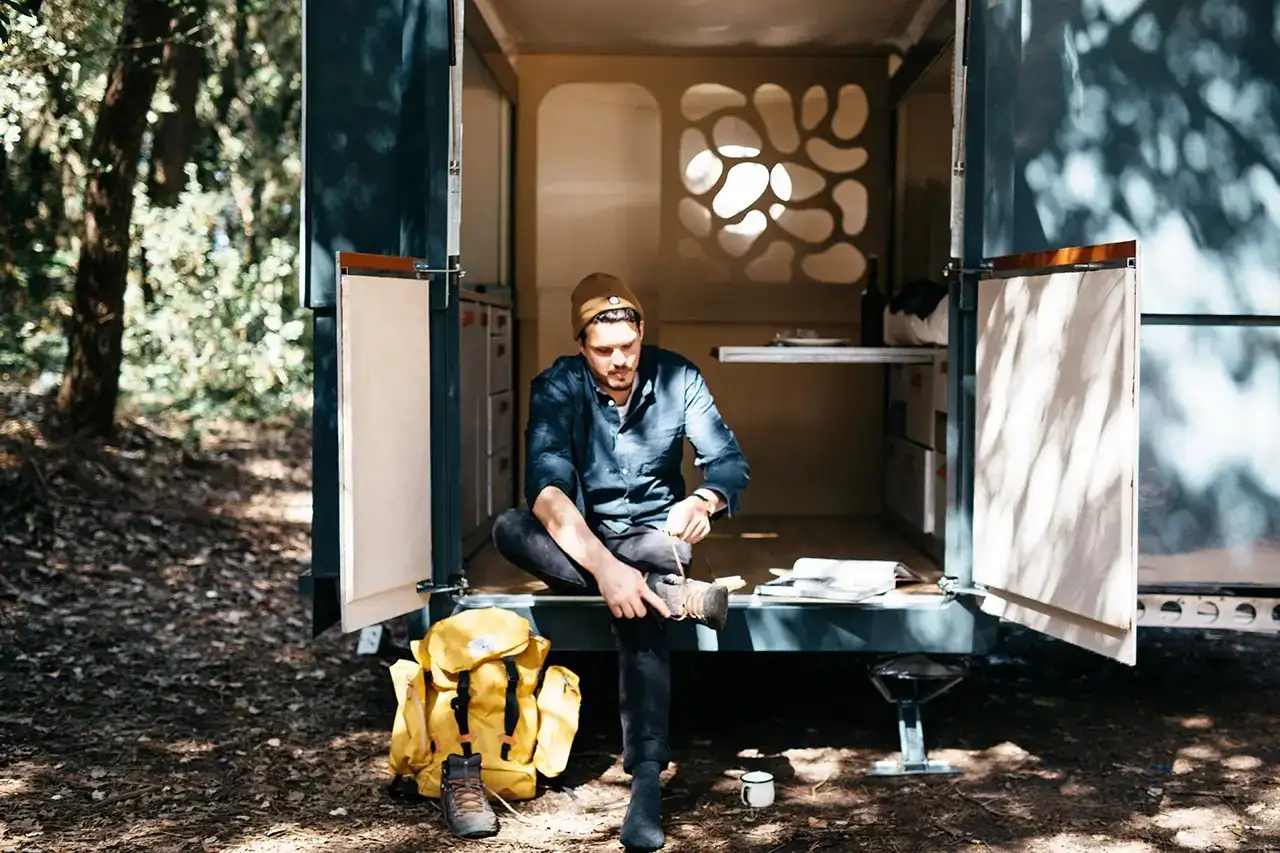 Person sitting at the back of a camper with a backpack and boots in a forest setting in British Columbia, Canada
