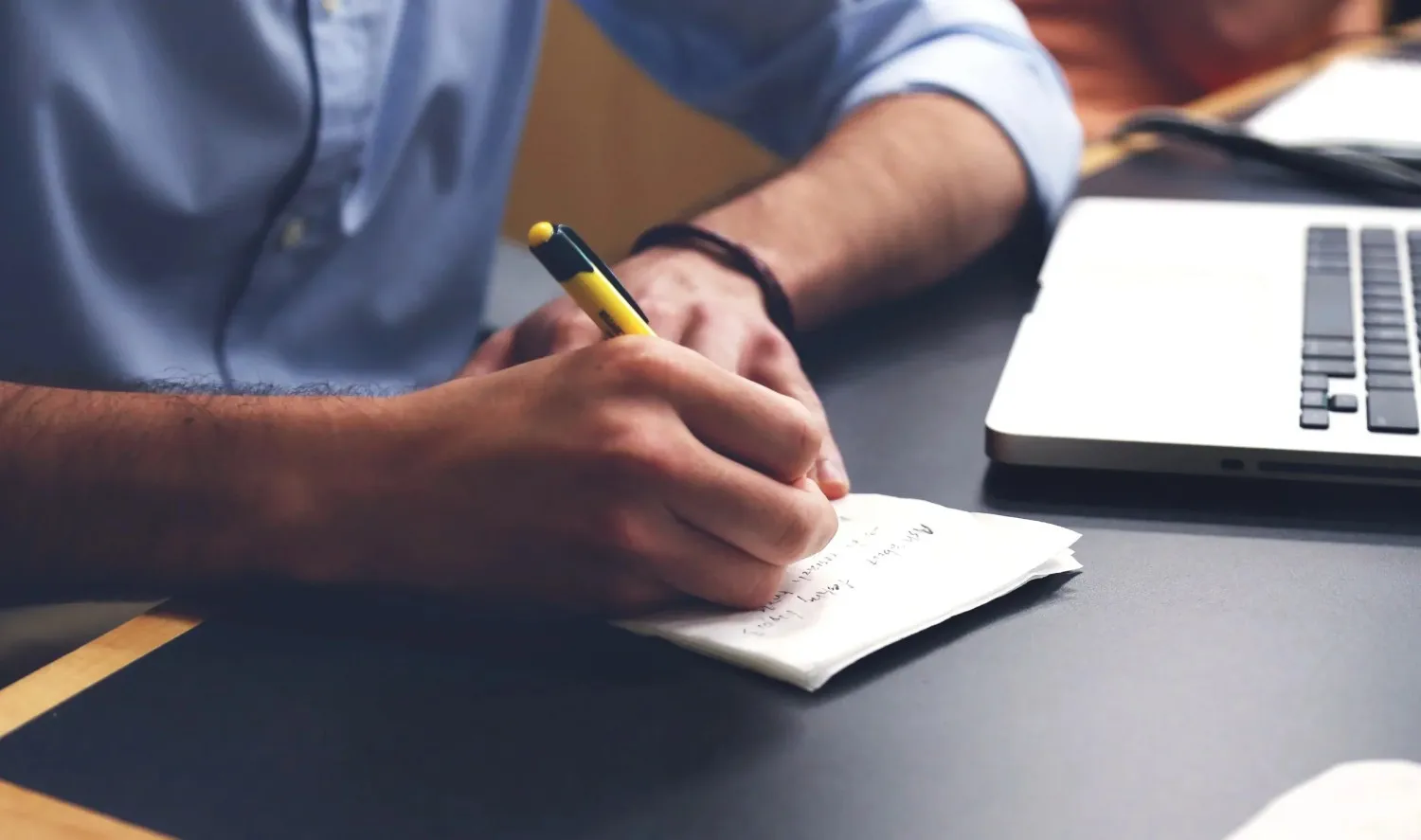 Person writing notes beside an open laptop at a desk in British Columbia, Canada